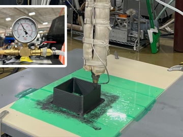 A 3D printing nozzle wrapped in insulation extrudes black composite material into a small square mold on a green and white flat surface in a lab setting. Inset shows a close-up of a pressure gauge connected to brass valves and tubing.