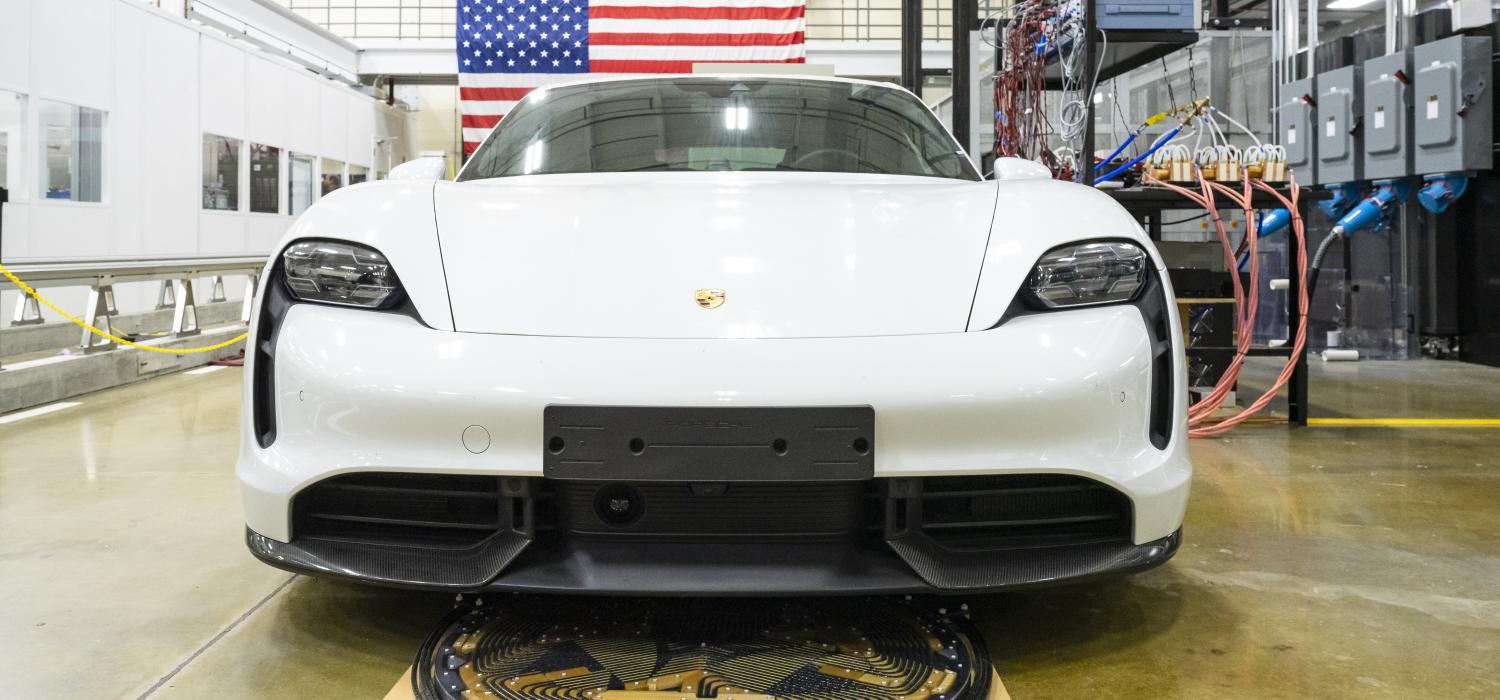 White sportscar parked almost over a wireless charging node of coils ont eh ground, wires and an American flag in background