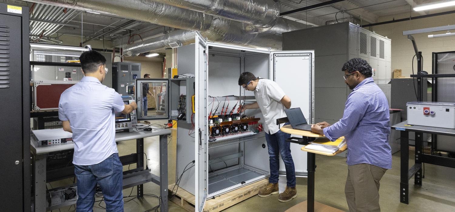 Three researchers working in a lab with large metal box open wand wiring visible inside