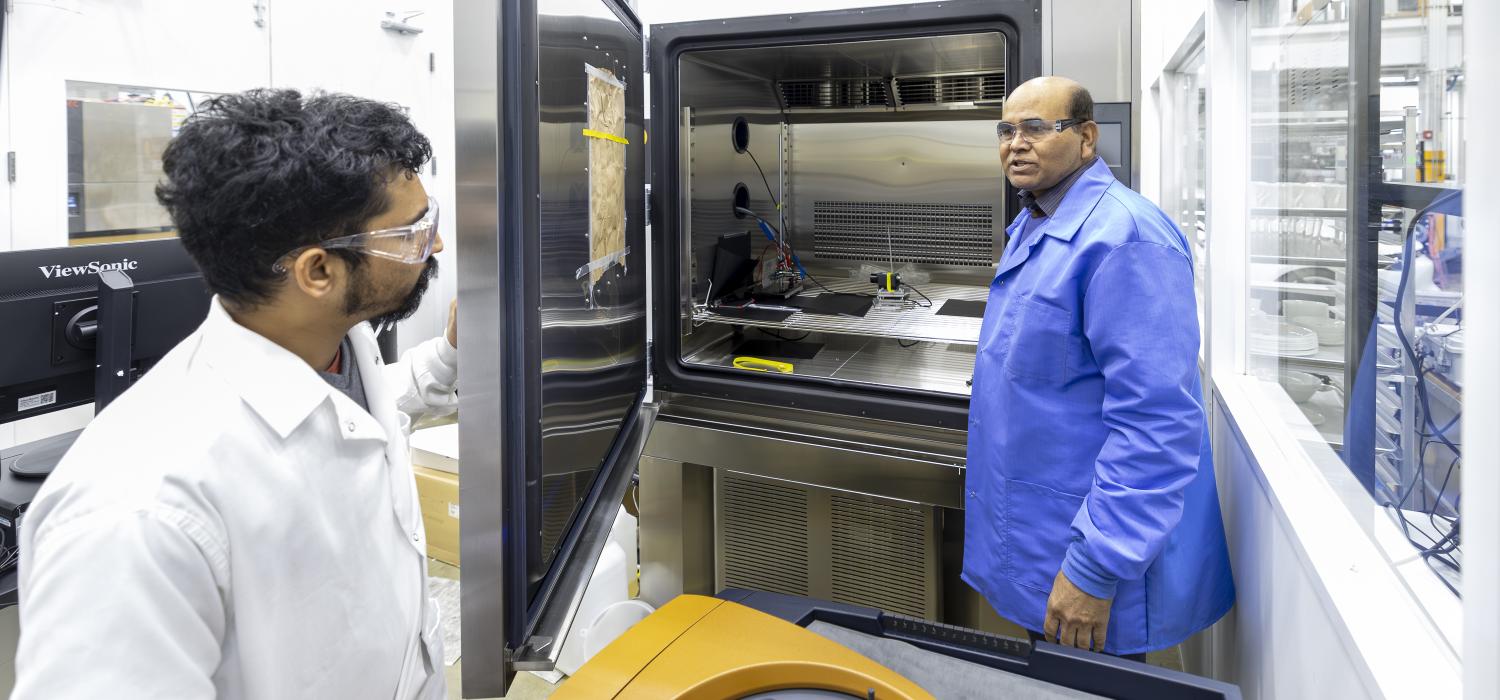 two researchers in lab coats next to temperature-controlled battery cycling machine that looks like a large stainless steel refrigerator with open door