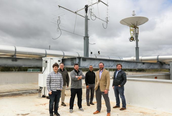 Six men pose in front of a satellite dish and antenna. 
