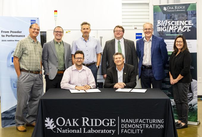 Eight people posing behind a table with a black tablecloth that says "Oak Ridge National Laboratory Manufacturing Demonstration Facility". There are papers and pens on the table