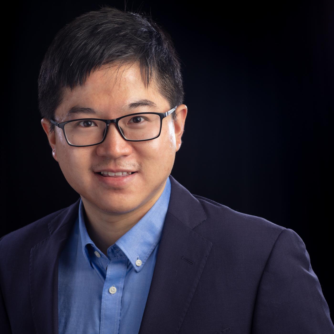 Man in blue suit jacket, blue button down shirt, glasses and dark hair smiles with a black backdrop. 