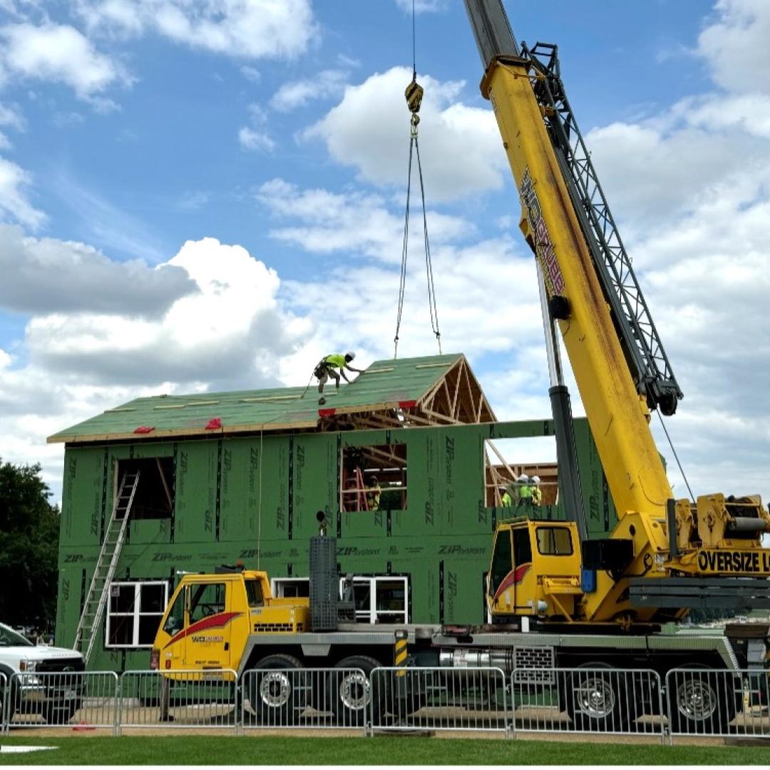 Green, two-story house is being assembled with the help of a yellow crane. 