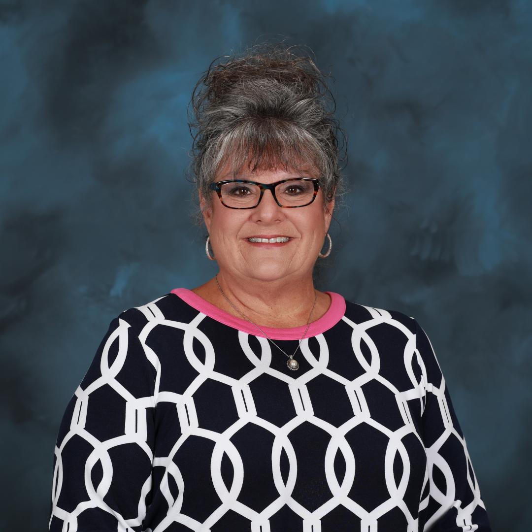 Image is of a woman with grey hair in an updo with black rimmed glasses and a black shirt with white circles and a pink neckline. 