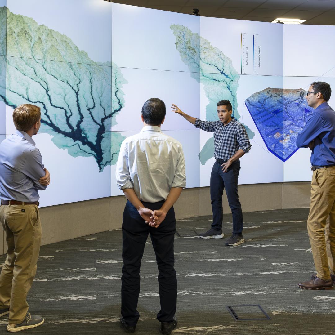This photo is of four men standing in front of a wall of monitors that are showing a tree looking image.