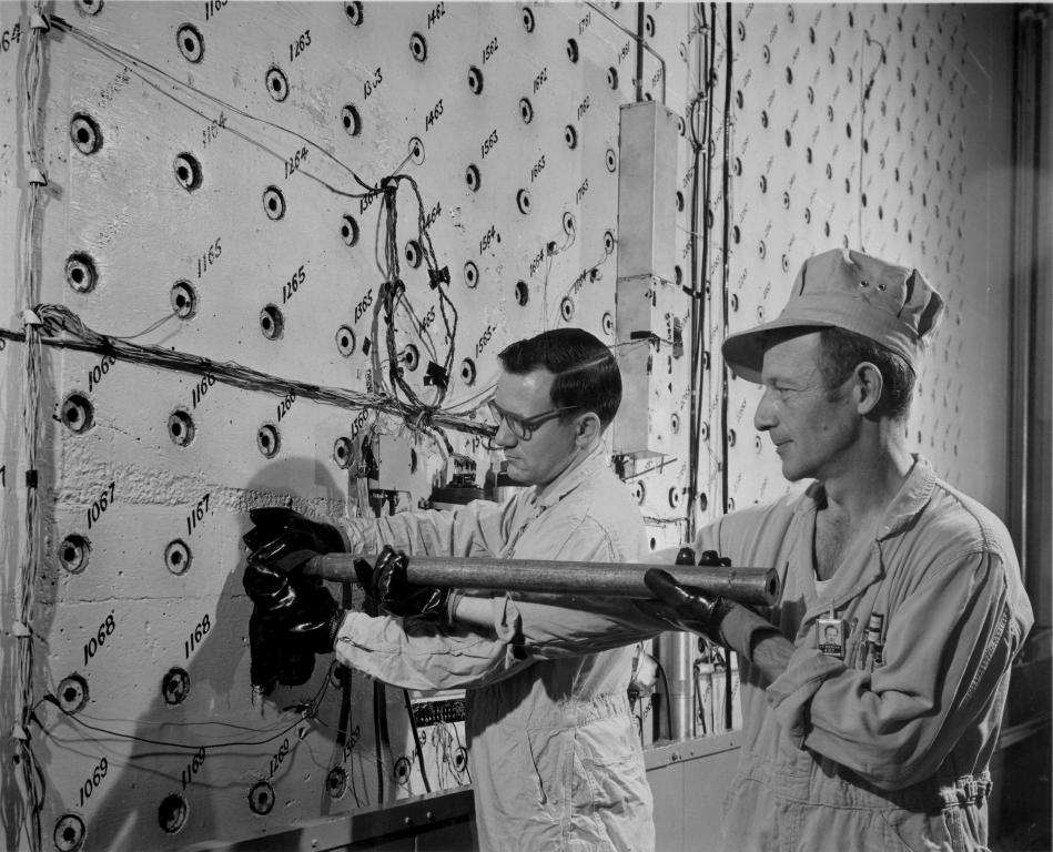 Two gentlemen using a push rod working at the Graphite Reactor loading face.