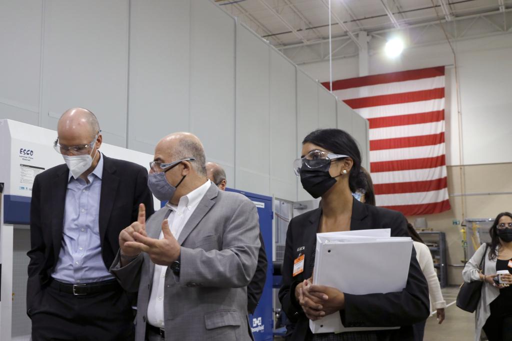 Several people walk and talk through a high-bay manufacturing facility, with an American flag in the background