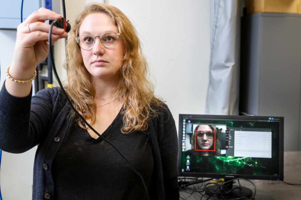 A woman looks into a handheld camera, which displays facial recognition on a monitor behind her