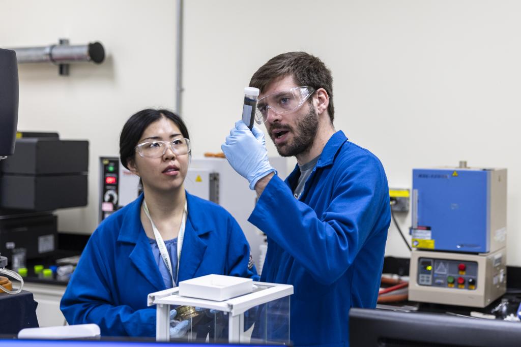 Two scientists in blue lab coats in battery lab, one holding a sample up while they discuss it