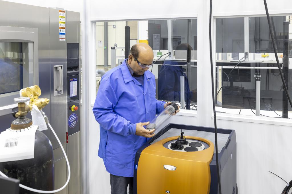 Researcher in blue lab coat holding glass cylinder next to square orange machine that measures heat from charging or discharging batteries