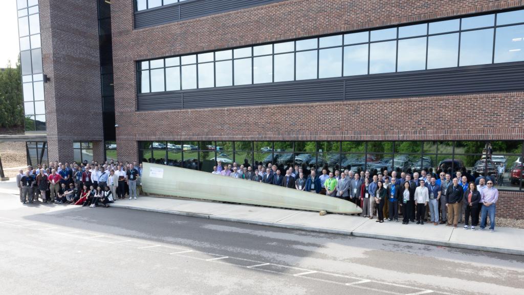 Group of more than 20 people standing in front of a brown building with a wind turbine tip that is long, white and looks like an elongated triangle.  