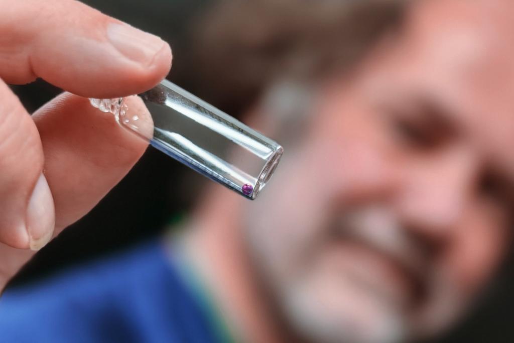 Man in a beard holding tweezers, showing a bead if space glass closer to the screen. 