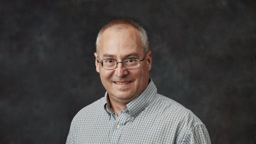 Man in glasses and a collared shirt smiles in front of a black backdrop