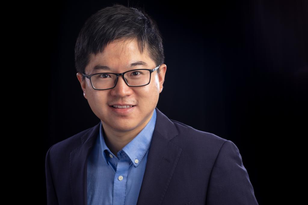 Man in blue suit jacket, blue button down shirt, glasses and dark hair smiles with a black backdrop. 