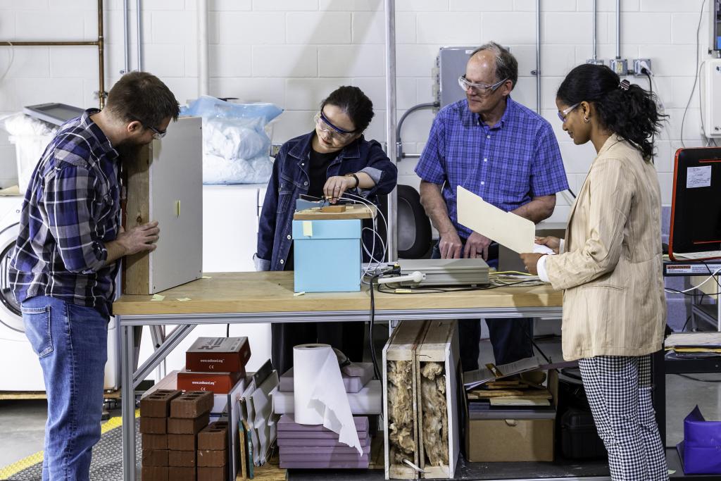 Four people stand around a table looking at a demonstration of a microwave moisture detector, a square grey material. 