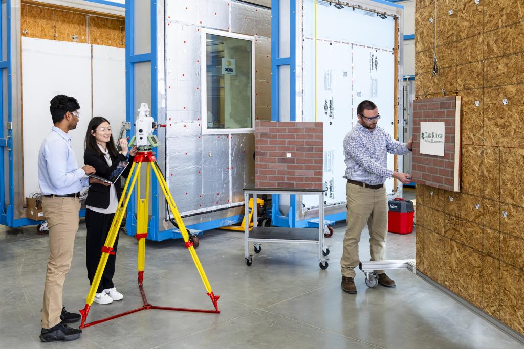 A man and a woman, left, are watching a man, right, hold up building material samples, such as a brick wall square to indicate alignment. 