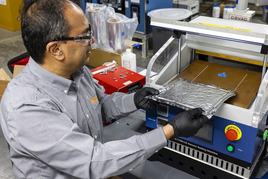 Scientist in goggles is wearing black gloves holding a rectangular piece of foil next to a machine. 