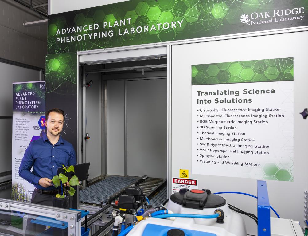 Man in blue shirt standing to the left of the screen is posted next to a sign that says advanced plant phenotyping laboratory, holding a plant.