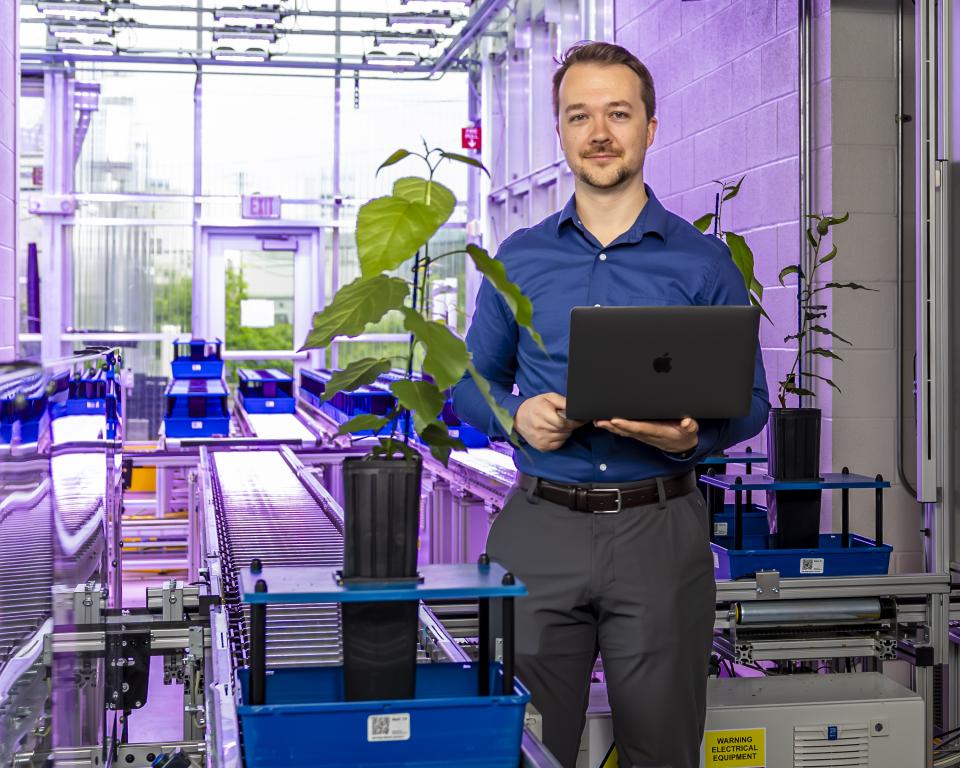 Man in blue shirt and grey pants holds laptop and poses next to a green plant in a lab. 