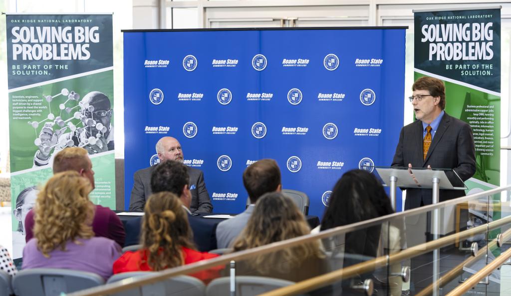 Speaker at the podium with a blue backdrop and two banners addresses group of people.