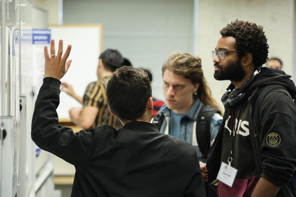 three students stand in front of a poster board with one explaining to the others his presentation