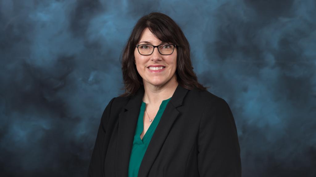 Woman with dark brown hair, glasses wearing a green shirt underneath a black blazer jacket smiling for a photo in front of blue backdrop