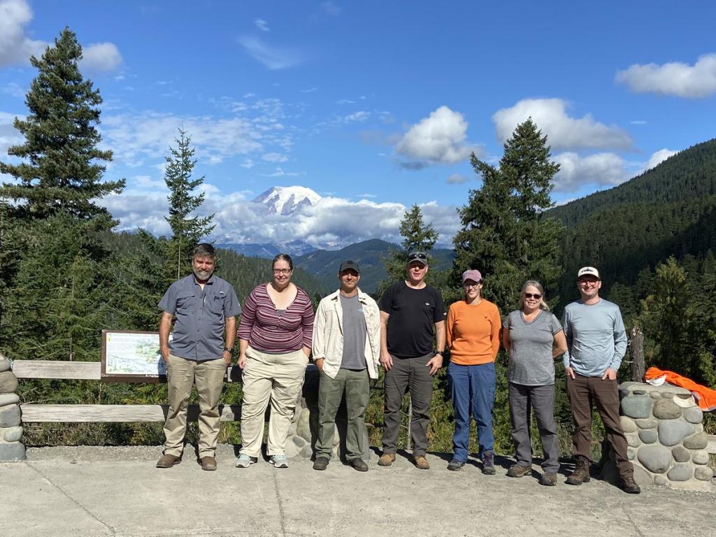 7 people are standing in a row in front of a mountain background, snow covered caps and green pines frame the photo