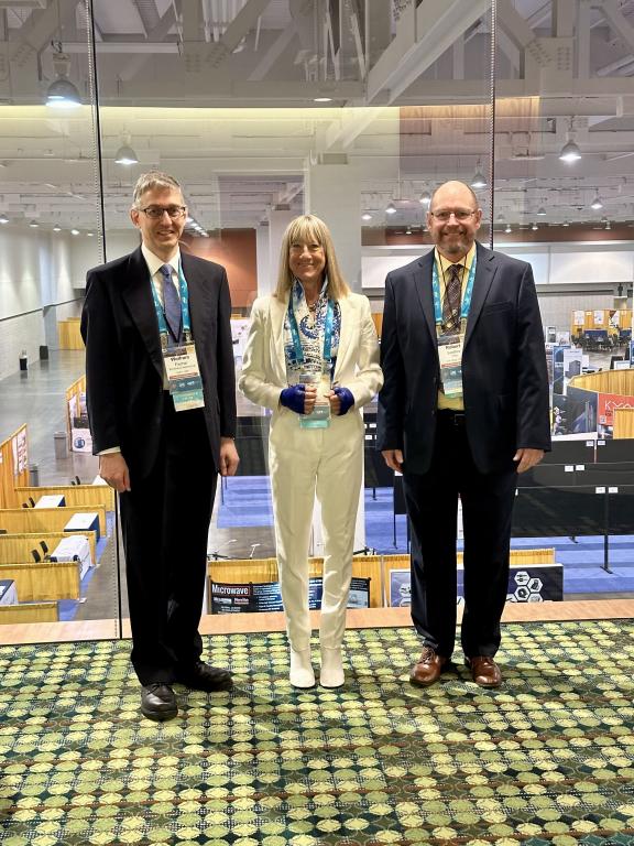 Two men and a woman stand on bright green carpet dressed in suites in the exhibit hall.