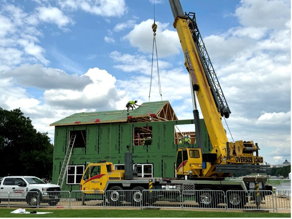 Green, two-story house is being assembled with the help of a yellow crane. 