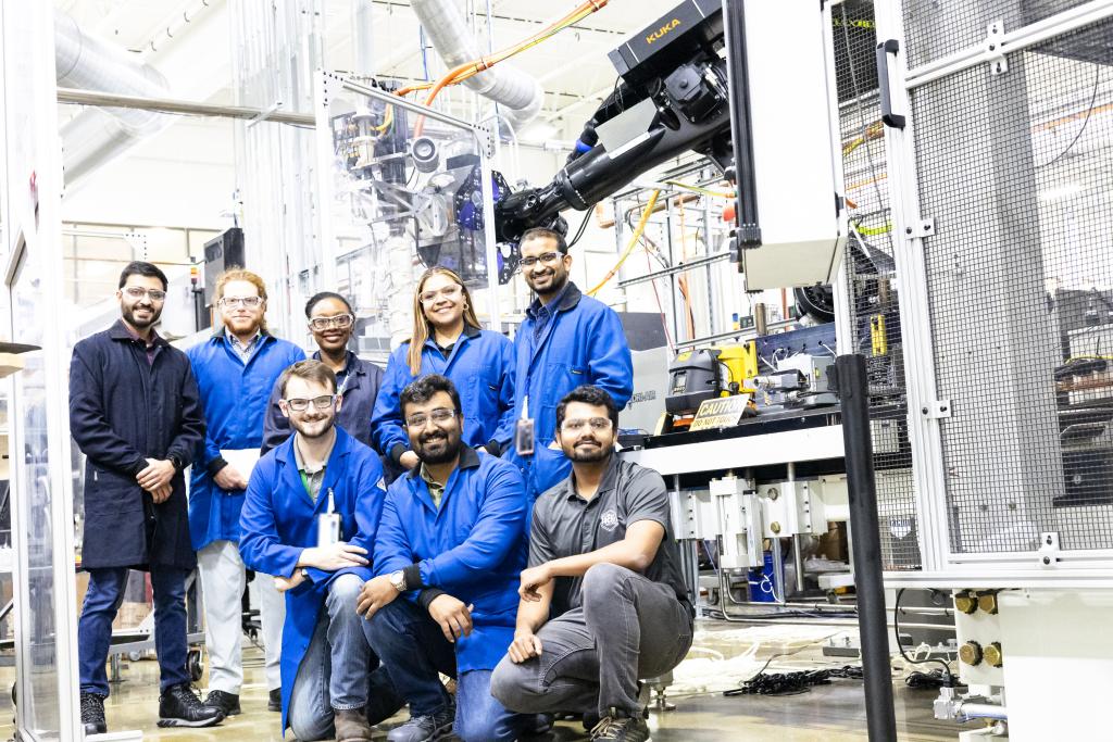 Six students in blue lab coats pose in industrial setting with two researchers in black lab coats
