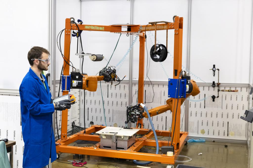 A person in a blue lab coat wearing gloves holds a control device in front of an orange machine