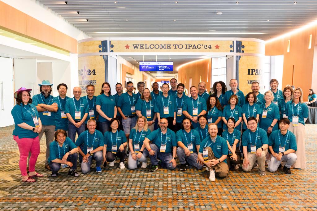 Big group of adults pose in teal colored tshirts under the Welcome to IPAC'24 sign. 