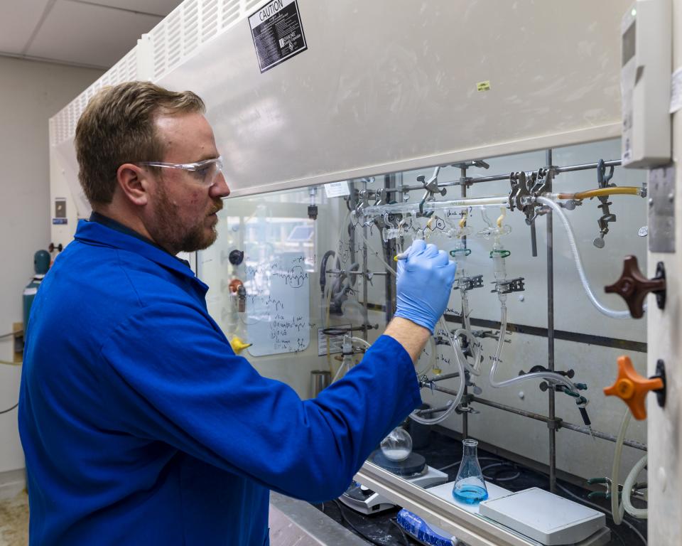 Man in a blue lab coat, gloves and glasses is working with materials in the lab