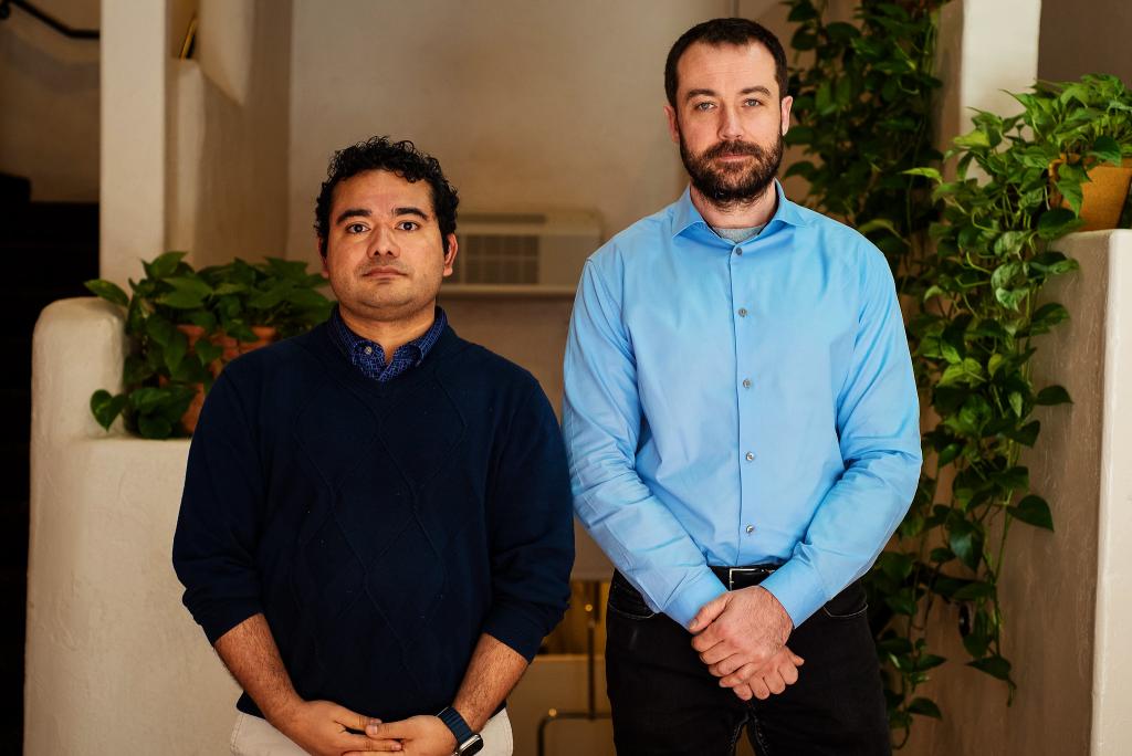 Two men are standing next to eachother posting for a photo. The man on the left is wearing a black long sleeve and the man on the right is in a bright blue button down