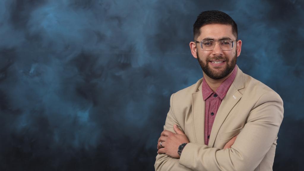 Man in tan jacket, red button down and glasses is smiling for a photo. He has short brown hair and his arms are crossed. 