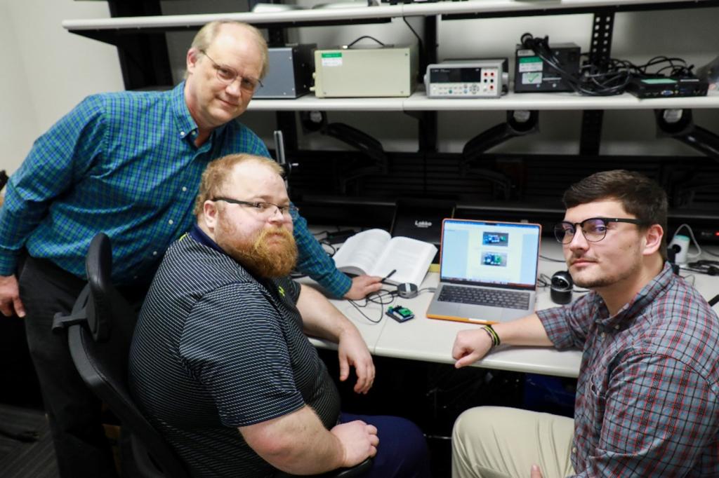 This photo is of three men sitting around a laptop computer that happens to be working on cybersecurity testing equipment.