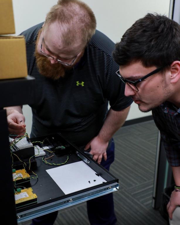 This picture shows two men looking down at technology on a table, with the one on the lefthand side holding a cord. 