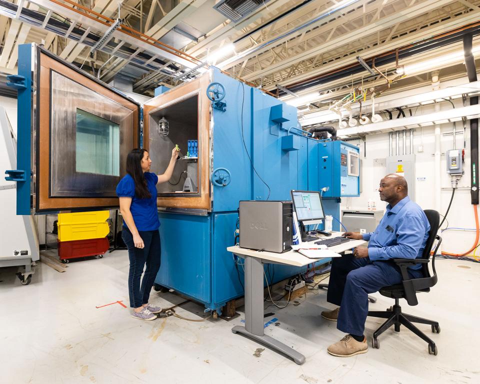 A woman places an object in a large blue metal testing chamber, while a man sits at a laptop next to it