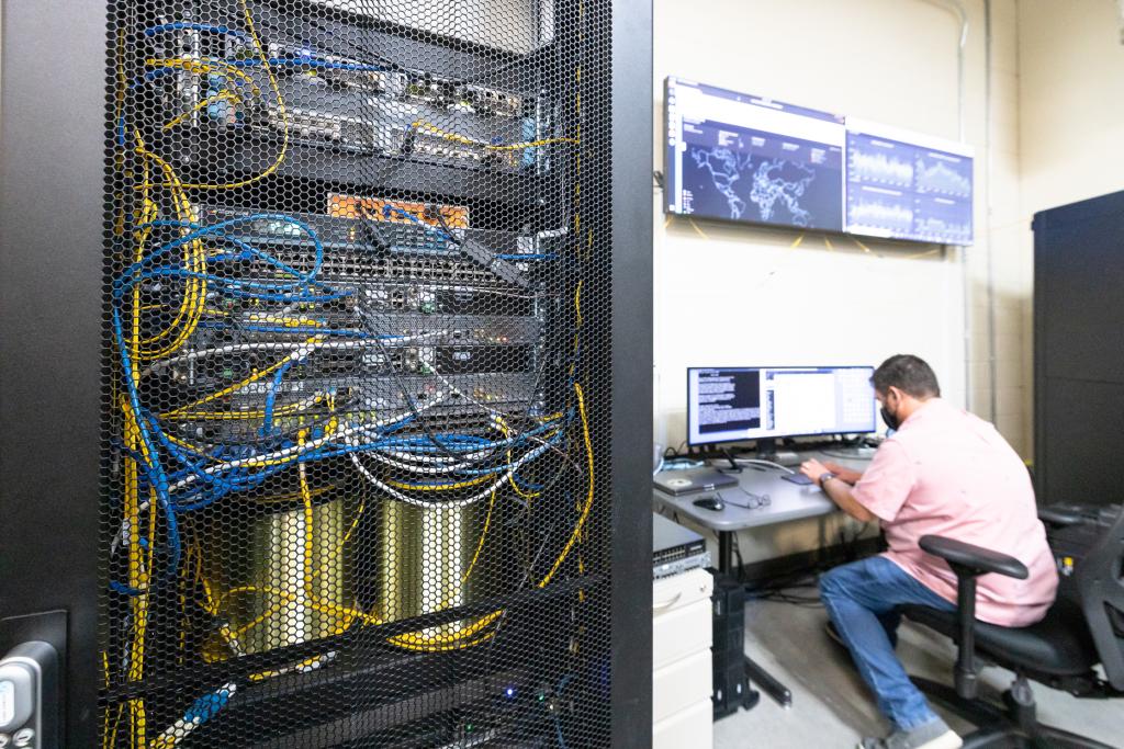 cabinet of wires and electronics behind mesh in foreground, man working at laptop in background