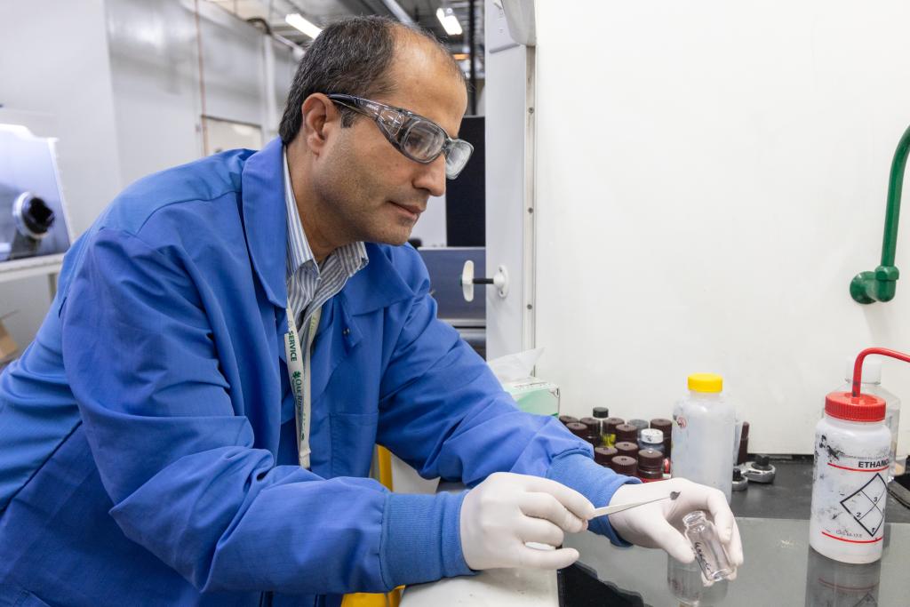 Man in blue lab coat moving material in into a vessel using tweezers in a lab