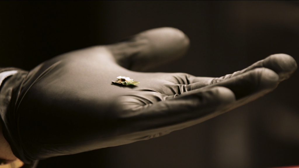 A hand in a black glove holds a heater platform designed to deliver atomized material to a sample