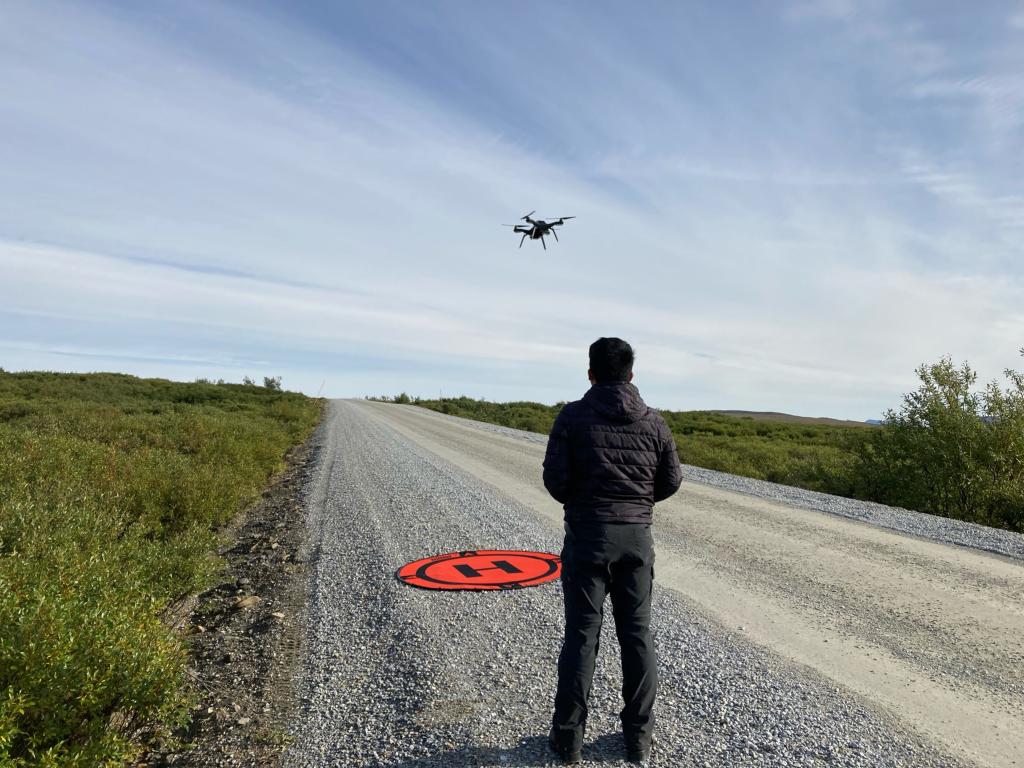 Daryl facing away from the camera flying a drone over a gravel road under a blue sky