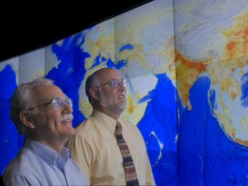 Two men look at a world map displayed on a large screen