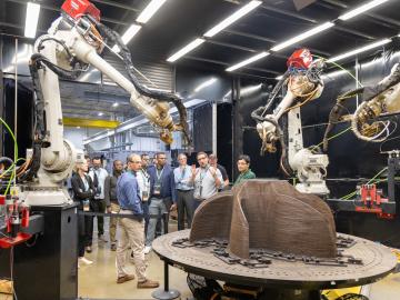 Two robotic arms, one on the left side of the frame and one on the right - are pointing at a brown three pronged shape on a table. There are people in the background looking at this setup.