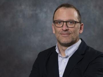 Man in suit jacket and glasses poses for photo in a grey background.