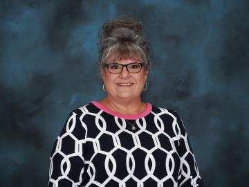 Image is of a woman with grey hair in an updo with black rimmed glasses and a black shirt with white circles and a pink neckline.