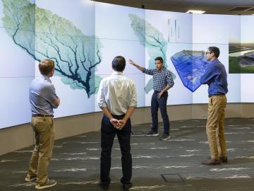 This photo is of four men standing in front of a wall of monitors that are showing a tree looking image.