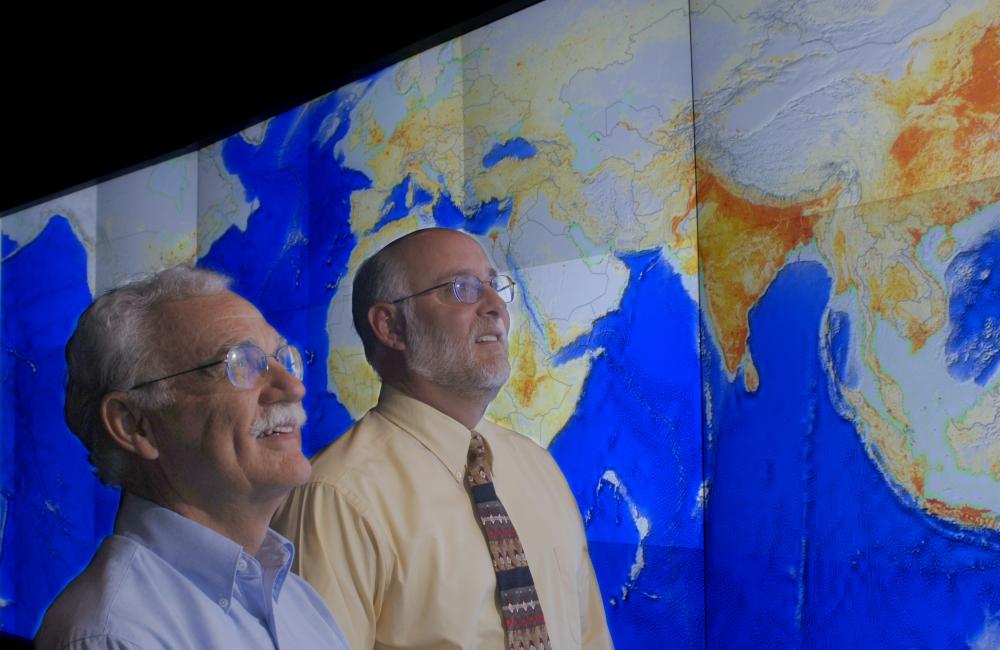 Two men look at a world map displayed on a large screen
