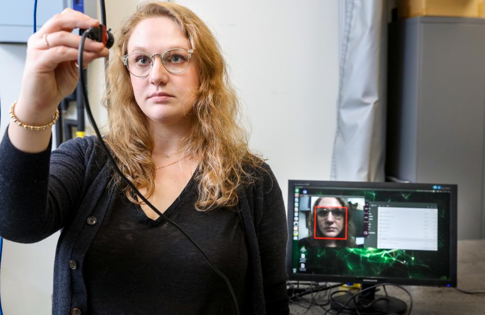 A woman looks into a handheld camera, which displays facial recognition on a monitor behind her